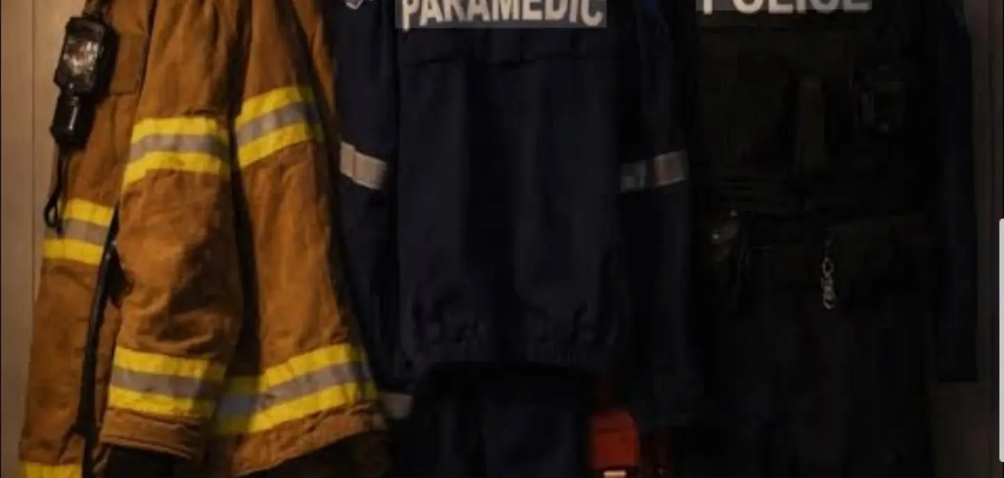 A firefighter's helmet and turnout coat, a paramedic jacket and a police uniform hanging side by side on a wall